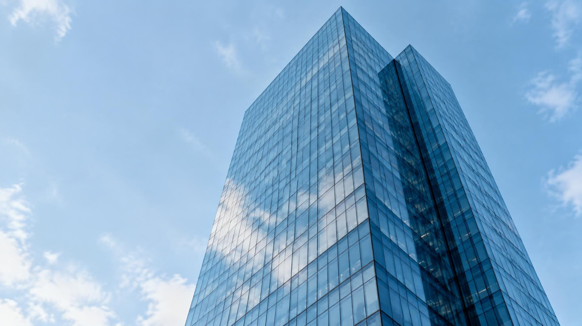 Modern glass office tower seen from below against a soft blue sky